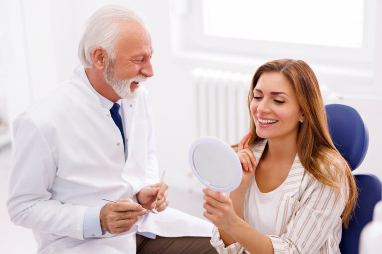 Woman looking in the mirror and smiling after checkup at dentist office; dentist and patient at dental clinic