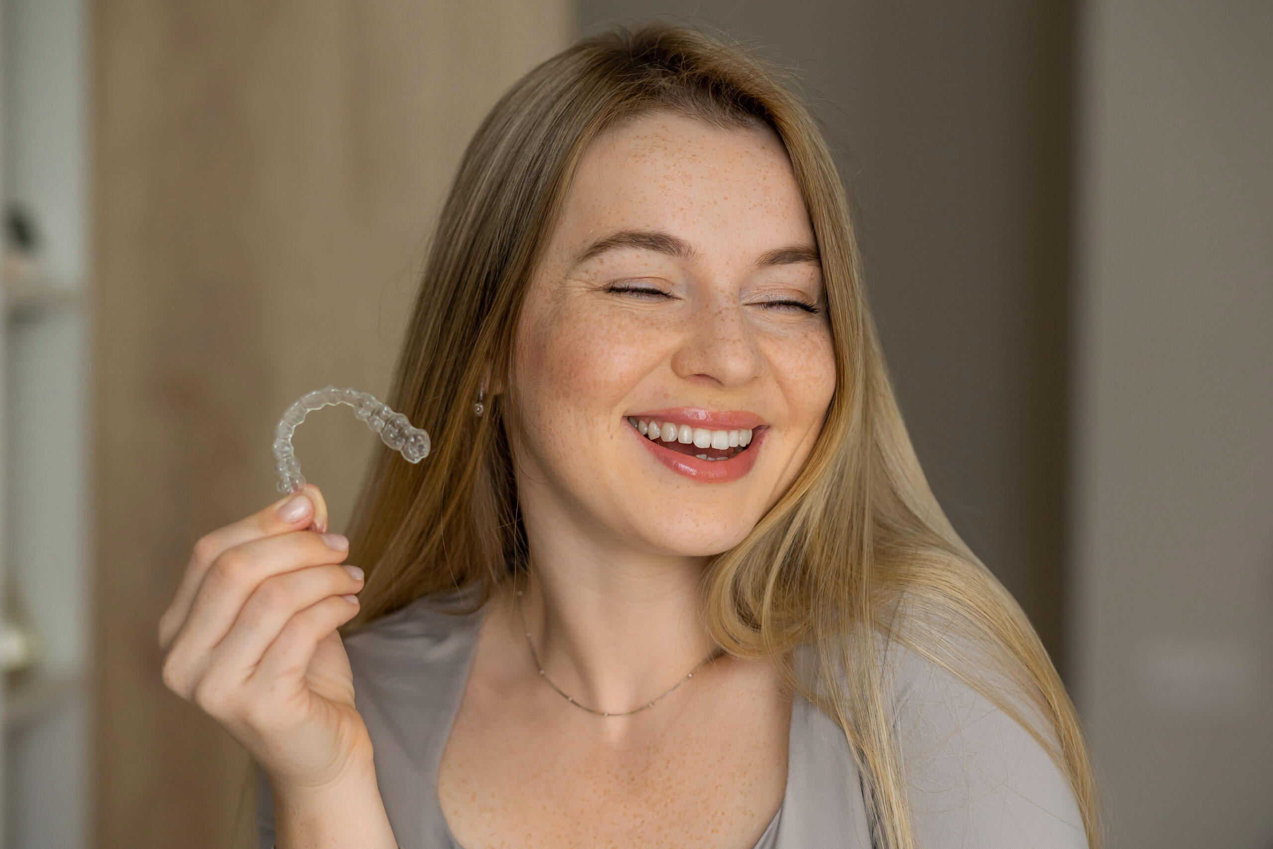 A cheerful young woman is smiling while holding a dental aligner for orthodontic treatment.